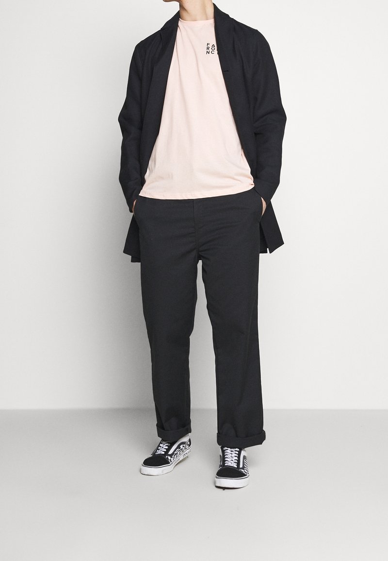 Veste longue noire, t-shirt rose avec texte, pantalon noir et chaussures noir et blanc. Design simple, coupe décontractée et look casual.
