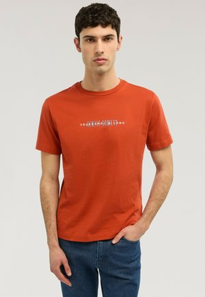 Junger Mann mit kurzen lockigen Haaren, der ein oranges T-Shirt mit rundem Ausschnitt und weißem Text sowie eine blaue Jeans trägt, vor einem einfarbigen Hintergrund steht.