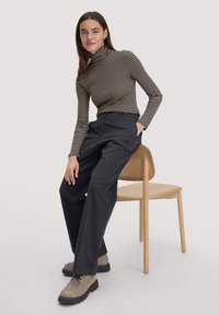 Gestreepte coltrui in zwart en beige, gecombineerd met hoge, donkere broeken. Het model zit op een lichthouten stoel en draagt taupe veterschoenen.