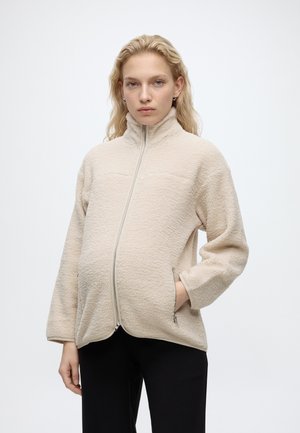 Femme aux cheveux blond clair portant une veste polaire beige zippée et un pantalon noir, debout avec une main dans la poche devant un fond uni.