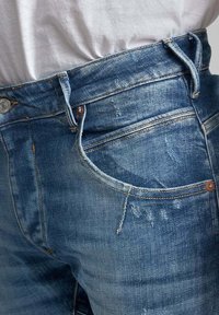 Jeans en denim bleu avec une coupe ajustée, une texture légèrement décolorée, un bouton en métal et des accents de couture sur la poche et la ceinture.