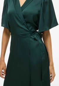 Smaragdgrønn sateng omslagskjole med korte volangsleeves, med knyttet midje og glatt tekstur. Rene linjer fremhever designet.