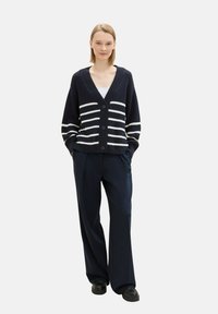 Marineblauer Strickcardigan mit weißen horizontalen Streifen, V-Ausschnitt-Design und drei Knöpfen. Kombiniert mit dunklen Hosen und schwarzem Schuhwerk.