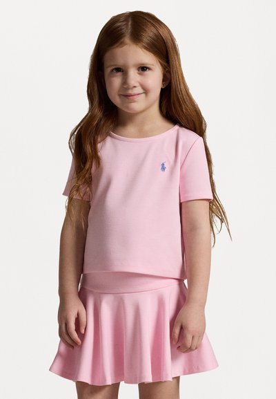 Svetlo roza majica s kratkimi rokavi in ujemajoča se krila v zvonu. Mehka bombažna tkanina z modrim logotipom polo na prsih. Gladka tekstura in preprost dizajn.