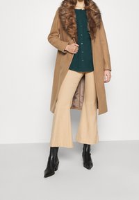 Manteau long en camel avec col en fourrure, chemise verte à boutons, et pantalon beige évasé ; bottines à talons noires ; textures lisses et couleurs neutres.