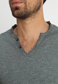 Mężczyzna z zarostem, ubrany w szary t-shirt henley z dwoma ciemnymi guzikami rozpiętymi, odsłaniającym owłosienie na klatce piersiowej i luźno zapiętą kołnierzyk.