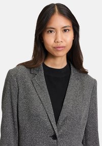 Szary marynarka o teksturowanej powierzchni, z klapami, z pojedynczym zapięciem na guzik i subtelnym wzorem w jodełkę. Nosi się ją na czarnym swetrze.