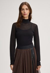 Maglia nera a maglia con maniche lunghe, dotata di collo alto e motivo a nido d'ape. Indossata con una gonna trasparente marrone, caratterizzata da un cinturino con logo.
