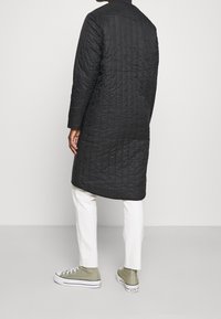 Abrigo negro acolchado con cuello alto, mangas largas y corte a media longitud, combinado con pantalones blancos y zapatillas altas grises.