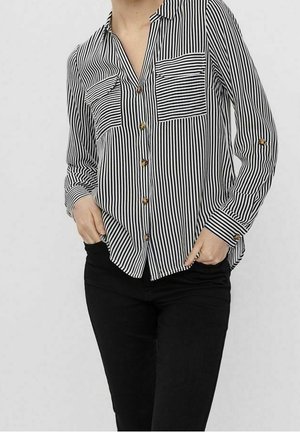 Kobieta ubrana w czarną i białą koszulę zapinaną na guziki w pionowe paski, z kieszenią o paskach ułożonych poziomo, w parze z czarnymi spodniami, ręce w kieszeniach.