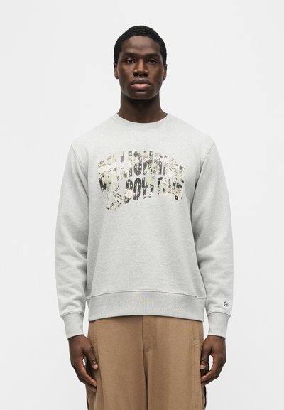 Άνδρας που φοράει ανοιχτό γκρι φούτερ με μοτίβο κειμένου "California Sports" και μπεζ παντελόνι, στέκεται μπροστά σε απλό λευκό φόντο.