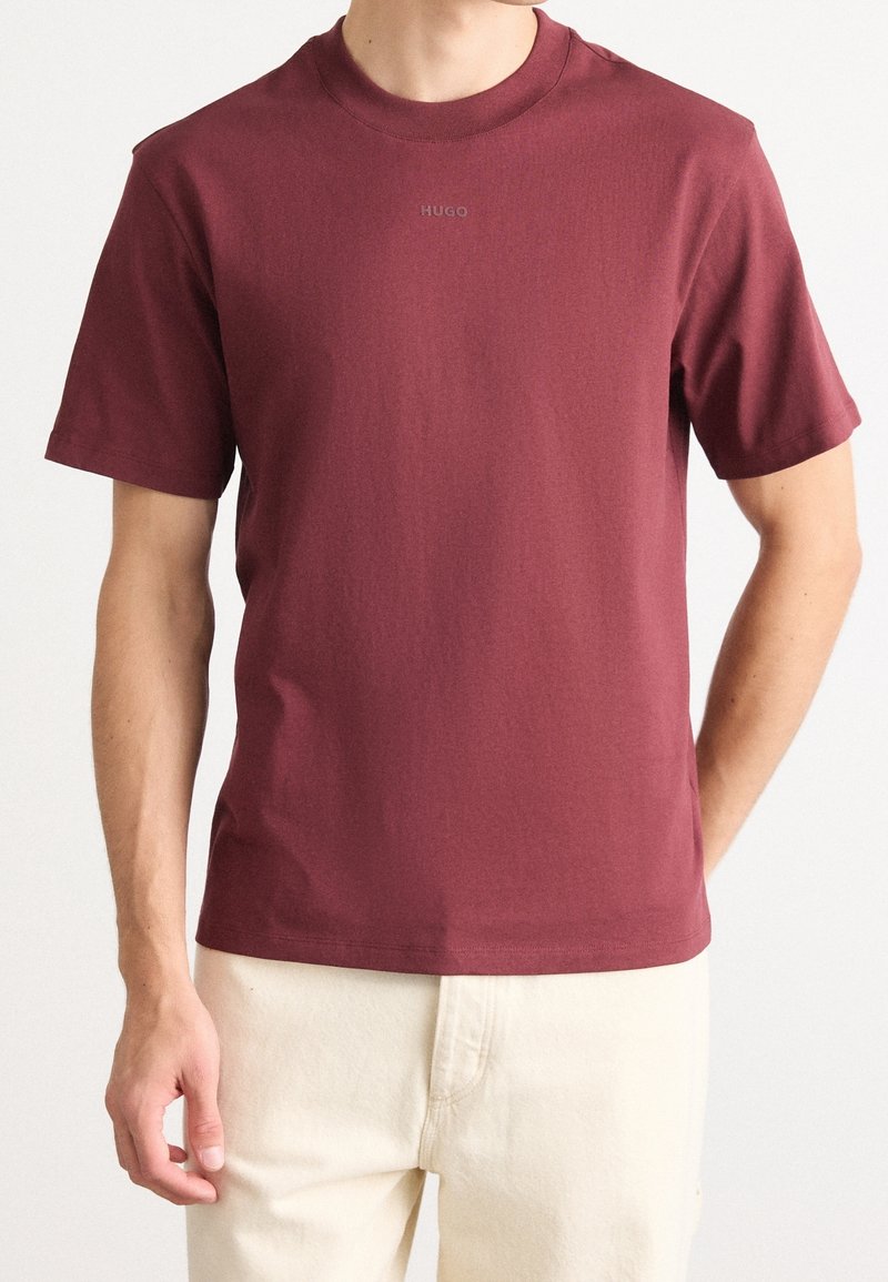 Un homme porte un T-shirt ajusté à manches courtes de couleur bordeaux avec le logo "HUGO" sur la poitrine et un pantalon blanc cassé, sur un fond clair uni.