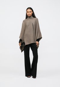 Hnědý pletený pončo s vysokým límcem a předním rozparkem, doplněný černými širokými kalhotami. Materiál působí měkce s texturovaným povrchem.
