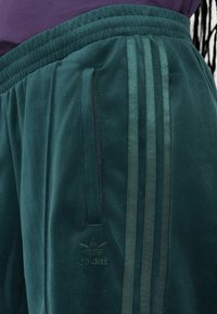 Zelené velúrové jogger nohavice majú tri tónové pruhy po stranách, pás s šnúrkou a malé embosované logo na ľavom stehne.