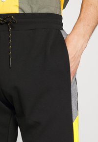 Svarta sweatpants med gula, grå och svarta paneler. Har en elastisk midja med snörning och sidofickor. Tillverkad av mjukt tyg.