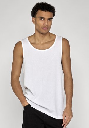 Jonge man met krullend haar, gekleed in een wit mouwloos hemd en zwarte broek, staand met één hand in de zak tegen een effen achtergrond.
