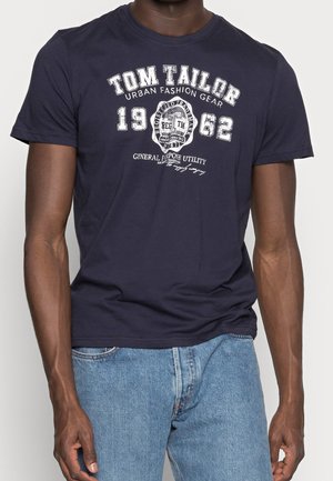 Homme portant un t-shirt graphique Tom Tailor bleu marine et un jean bleu clair, se tenant contre un fond blanc uni.