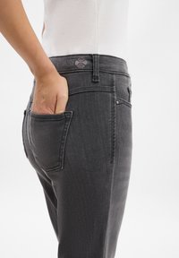 Mano insertada en el bolsillo trasero de unos vaqueros negros desgastados, combinados con una blusa blanca metida por dentro, presentado sobre un fondo blanco liso.