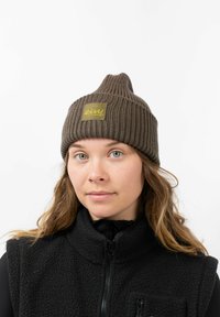 Brown ribbed knit beanie with a peak, featuring a square woven patch with the brand name. Worn with a black fleece vest.