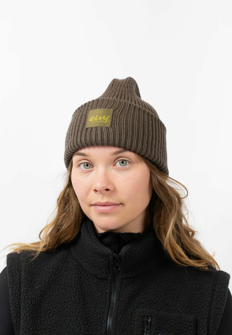 Brown ribbed knit beanie with a peak, featuring a square woven patch with the brand name. Worn with a black fleece vest.