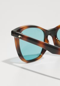 Occhiali da sole tartarugati con lenti turchesi e design arrotondato. Presenta un accento dorato sulla tempia. Montatura in plastica liscia e lucida.