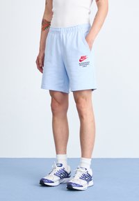 Person som bär ljusblå Nike-shorts med texten "Endast auktoriserad personal", vit linne, vita strumpor och vit-lila Nike-sneakers.