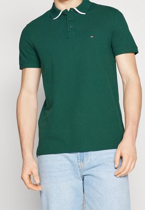Mann trägt ein dunkelgrünes kurzärmliges Poloshirt mit weißer Einfassung am Kragen und hellblaue Jeans, steht mit entspannten Händen.