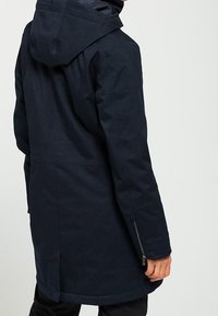 Navy blue parka jacket with a textured fabric, hood, and zippered side pockets. Straight cut design with a slightly longer back hem.