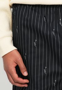 Pantalons à rayures noires avec de fines lignes blanches et des accents abstraits blancs sporadiques. Le matériau semble lisse, ajusté à la taille, avec une manche crème texturée visible.