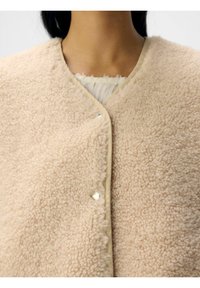 Beige faux shearling vest med blød, tekstureret overflade og rund krave. Lukket med en trykknap foran, med kontrastkant.