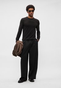 Černý pletený svetr, černé široké kalhoty a hnědá semišová bunda přehozená přes jeden rameno. Model nosí tmavé sluneční brýle a černé boty.