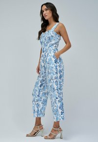 Mono floral en azul y blanco, con piernas anchas, tirantes ajustables, dos bolsillos y un cuerpo ajustado con detalles de botones.