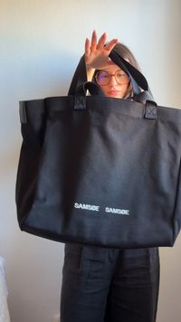 Une personne portant des lunettes tient un grand sac cabas noir SAMSØE SAMSØE, cachant la majeure partie de son visage et de son torse devant un mur uni.