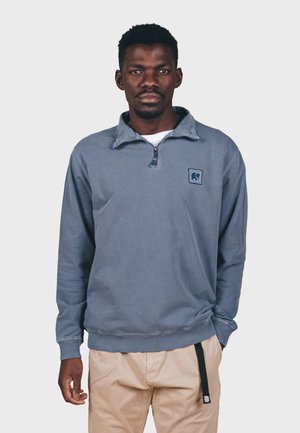 Pull bleu clair en polaire avec un demi-zip et un col. Présente un écusson logo sur la poitrine et des poignets côtelés, associé à un pantalon beige.