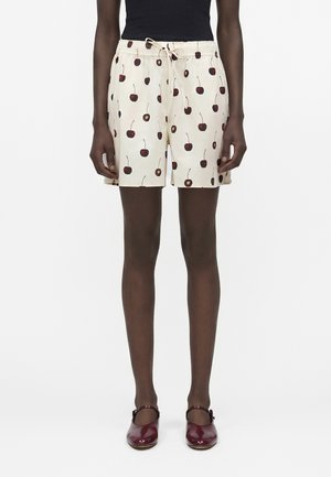 Személy krémszínű rövidnadrágot visel piros cseresznye mintával, fekete felsővel és sötétvörös Mary Jane cipővel, fehér háttér előtt áll.