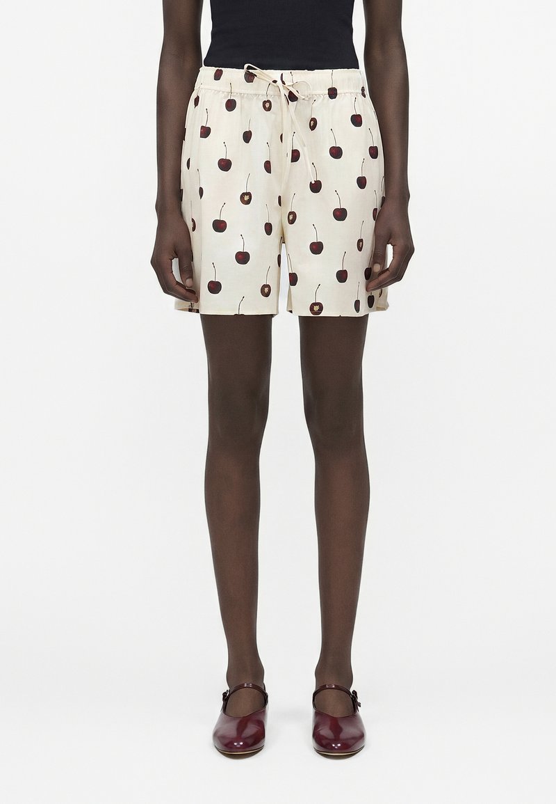 Άτομο που φοράει κρεμ σορτς με κόκκινο τύπωμα κερασιού, μαύρο τοπ και σκούρα κόκκινα παπούτσια Mary Jane, στέκεται μπροστά σε λευκό φόντο.