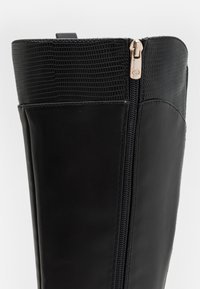 Botte noire montante jusqu'au genou avec une partie inférieure en simili cuir lisse et une partie supérieure en simili cuir texturé. Fermeture zippée sur le côté avec des accessoires en métal doré.