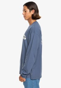 Koszulka z długim rękawem typu crewneck w niebieskim kolorze, wykonana z bawełny, z grafiką z logo z przodu, luźny krój i boczne rozporki na dole.