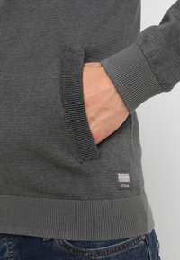 Main dans la poche d'un pull gris en maille avec poignets et ourlet côtelés, associée à un jean en denim bleu, montrant la texture du tissu et l'étiquette du vêtement.