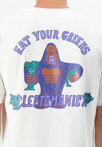 Weißes Baumwoll-T-Shirt mit einer bunten Grafik eines großen Wesens, das Brokkoli hält, mit dem Text "EAT YOUR GREENS" und "CLEPTOMANICY" in fetter Schrift.