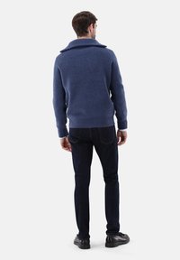 Blauer gestrickter Pullover mit weitem Kragen, geripptem Muster und langen Ärmeln, kombiniert mit dunklen Jeans und schwarzen Schuhen, von hinten betrachtet.