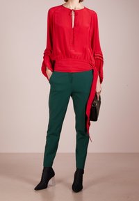 Červená hedvábná blůza s kulatým výstřihem a detaily na zavázání, v kombinaci s dokonale střiženými zelenými kalhotami. Černé texturované kotníkové boty a malá černá kabelka.