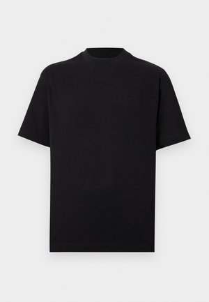 Ung mand står op ad hvid væg iført sort t-shirt, beige ternede knælange shorts, hvide sokker og sorte sneakers.