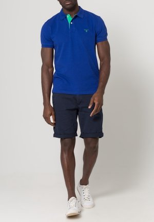 Blå pikétröja med grön krage, korta ärmar och en liten logotyp på bröstet, matchad med marinblå shorts och vita sneakers.