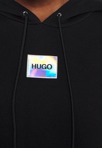 Svart huva sweatshirt med en glänsande holografisk rektangulär etikett med "HUGO" tryckt i fet svarta bokstäver. Enkelt design med snoddar.