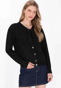 Zwarte cardigan met een gespikkeld patroon, ronde halslijn, voorste strikdetail en goudkleurige knopen. Draagt met een spijkerrok.