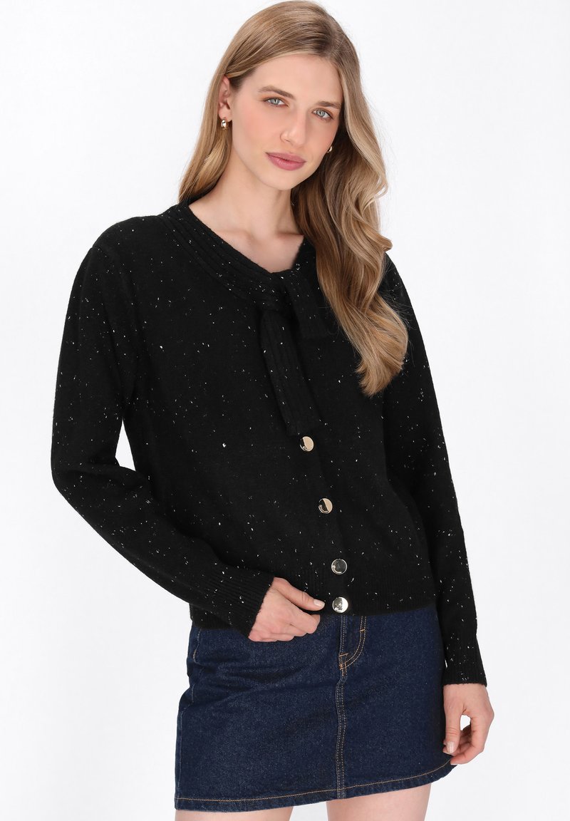 Zwarte cardigan met een gespikkeld patroon, ronde halslijn, voorste strikdetail en goudkleurige knopen. Draagt met een spijkerrok.