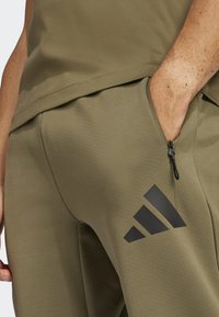 Calças de desporto verde-oliva com um acabamento texturizado, apresentando um bolso com fecho e um logótipo preto da Adidas na lateral.