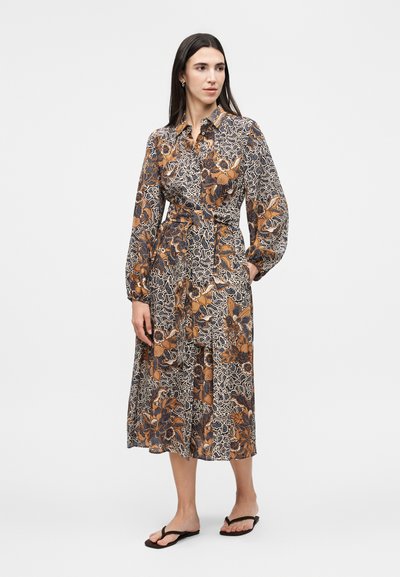 Γυναίκα με μακρυμάνικο φόρεμα μέχρι τον αστράγαλο με μπλε, πορτοκαλί και λευκά λουλουδάτα και αφηρημένα σχέδια, στέκεται με το ένα χέρι στην τσέπη.