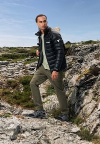 Sort pufferjakke, lysegrøn skjorte, olivenfarvede bukser, grå vandresko med sorte accenter. Udendørs miljø, klippeagtig terræn og grønne planter.